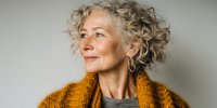 Elegant senior woman with curly gray hair wearing mustard yellow cardigan looking thoughtfully to side against neutral background.