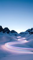 Light trails wind through snowy mountain valley at dusk, creating glowing pink and purple paths across pristine winter landscape with jagged peaks under clear blue sky.