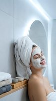 Young woman enjoying spa facial mask treatment in modern bathroom with towel wrapped hair relaxing during skincare routine at home wellness session.