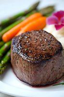 Juicy grilled beef tenderloin steak seasoned with salt and pepper, served with fresh asparagus, baby carrots and orchid flower garnish on white plate.
