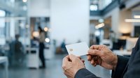 Close-up of two hands exchanging blank white business card in modern office lobby with blurred colleagues and glass architecture in background.