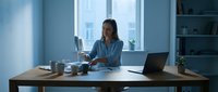 Young woman with dark hair and medium skin tone working at modern office desk with laptop, coffee cup, and notebook in bright contemporary workspace with large windows and shelving.
