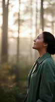 Woman with dark hair and East Asian ethnicity wearing a green jacket gazes upward in a misty forest surrounded by tall trees, conveying contemplation and connection with nature.