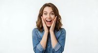 Young Caucasian woman with brown wavy hair showing excited surprised expression, hands on cheeks, wearing denim shirt on white background.