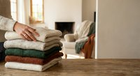 Woman organizing folded knitted sweaters stack on wooden table in cozy living room with fireplace. Home organization and seasonal clothing storage concept.