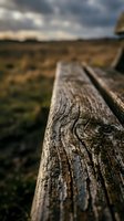 Weathered wooden fence rail in close-up with blurred rural landscape and dramatic sky in background, suitable for rustic and countryside design projects.