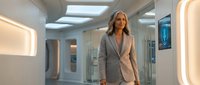 Middle-aged woman with long blonde hair, Caucasian ethnicity, wearing a gray blazer in a modern, minimalist corridor with futuristic lighting and glass partitions, suitable for corporate, business, and professional concepts.