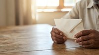 Person with dark skin tone holding white envelope at wooden table with warm natural light from window, suitable for business communication, mail delivery, correspondence, and office work concepts.