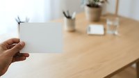 Woman holding blank white business card in modern office workspace with wooden desk, potted plants, and supplies in background.