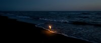 Solitary candle flame glowing on dark ocean beach at twilight with waves washing shore creating peaceful meditation atmosphere for relaxation and mindfulness.