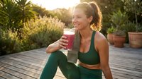 Young woman in green athletic wear holding a glass of pink smoothie, sitting on wooden deck surrounded by potted plants and greenery during golden hour sunlight, wellness and healthy lifestyle concept.
