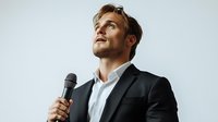 Young Caucasian male professional speaker in black suit jacket holding microphone, looking up with confident expression against light background, business presentation concept.
