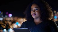 Woman with curly hair wearing black jacket using tablet at night with city lights bokeh background for technology and urban lifestyle content.