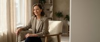 Professional middle-aged Caucasian businesswoman in beige suit sitting confidently in modern office interior with natural lighting and minimalist decor.