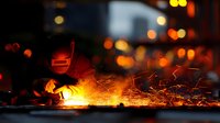 Welder in protective mask working with molten metal at night, creating dramatic shower of bright orange sparks against dark urban background.