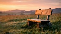 Wooden bench sitting alone in a misty meadow at sunrise, surrounded by rolling hills and mountains in soft golden light, perfect for contemplation and nature themes.