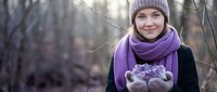 Young Caucasian woman in purple knit hat and scarf holding fresh mushrooms in forest during autumn foraging adventure outdoors in natural woodland setting.