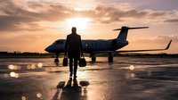 Silhouette of businessman walking towards private jet on airport tarmac during golden sunset with dramatic clouds and runway lights creating luxury travel scene.