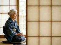 Senior woman in traditional dark kimono sitting seiza position on floor by shoji screen, holding tea cup, contemplative pose, peaceful indoor setting for wellness and cultural lifestyle content.