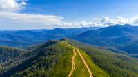 Aerial view of winding mountain road cutting through green ridge with dramatic blue mountain ranges and white clouds stretching to horizon on sunny day.