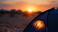 Illuminated camping tent glows against dramatic desert sunset with mountain silhouettes, creating warm atmospheric outdoor scene. Adventure travel concept.