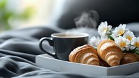 Freshly baked croissants on white plate with steaming coffee cup and delicate white flowers, moody dark background creates cozy breakfast atmosphere.