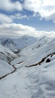 Snow-covered mountain peaks and valley under cloudy winter sky, with winding path through deep powder snow leading to dramatic alpine landscape views.