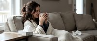 Sick woman with dark hair and light skin tone sitting on beige couch, holding tissue to face, appearing unwell with cold or flu symptoms at home.