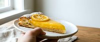 Slice of lemon tart with flaky pastry crust, glossy citrus curd filling, and candied lemon slices on a ceramic plate held by hand.