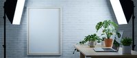 Blank wooden frame hanging on white brick wall above desk with potted plants, laptop, and professional lighting setup for modern workspace design.
