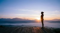 Woman in athletic wear standing on wooden platform overlooking misty valley at sunrise, contemplating mountain landscape during morning meditation or fitness journey.