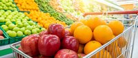 Red apples and oranges in shopping cart at grocery store with fresh produce display in background for supermarket shopping and healthy food retail concepts.