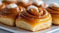 Fresh baked cinnamon rolls with caramel glaze and cream cheese frosting on white plate, close up view showing flaky pastry layers and gooey topping.