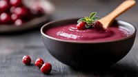 Fresh cranberry smoothie bowl garnished with mint and berries in black ceramic bowl, wooden spoon on dark background with scattered fruit.