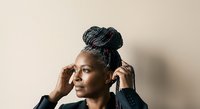 Mature Black woman with braided updo hairstyle wearing black blazer, adjusting hair with both hands against neutral beige background, professional portrait.