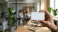 Close-up of hand holding blank white business card in modern office with blurred colleagues and plants in background, suitable for corporate branding and business identity mockups.