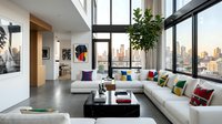 Modern loft living room with floor-to-ceiling windows overlooking city skyline, white sectional sofa with colorful pillows, black coffee table, potted plants, and contemporary artwork on walls.