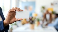 Male professional with dark skin tone holding blank white business card in office environment with blurred colleagues and workspace in background.