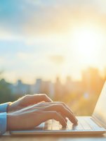 Close-up of hands typing on laptop keyboard against blurred city skyline at sunset, conveying remote work and digital lifestyle.