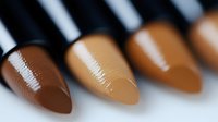 Macro shot of makeup foundation or concealer swatches in different skin tone shades showing creamy texture and glossy finish against blurred background.