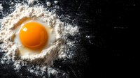 Raw egg yolk in white flour scattered on dark black surface, dramatic food photography with natural lighting and minimalist composition for culinary design.