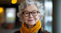 Smiling senior woman with curly white hair wearing round glasses and bright yellow scarf, expressing warmth and confidence against soft blurred background.