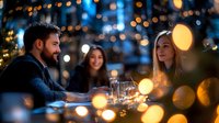 Young bearded man and two women enjoying evening drinks at outdoor cafe with festive bokeh lights creating warm atmosphere.