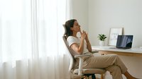 Young Asian businesswoman in casual attire sitting at modern home office desk, thinking thoughtfully with hand on chin near laptop computer in bright room.