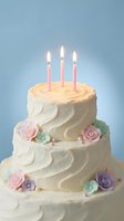 Three-tier white frosted birthday cake with pink candles and sugar flowers on blue background for celebration party dessert bakery design.
