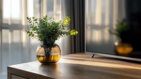 Green leaves in round amber glass vase on wooden table near window with soft natural light and shadows creating cozy morning atmosphere.