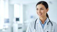 Young female doctor with stethoscope smiling confidently in modern medical office, healthcare professional ready for patient consultation.