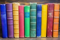 Row of colorful vintage hardcover books standing on wooden shelf, spines showing various sizes and textures against rustic background, arranged in rainbow order.