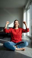 Happy young Caucasian woman in red sweater celebrating success with raised fists while sitting cross-legged on floor in modern apartment with natural light.