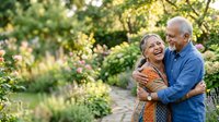 Senior couple embracing on garden path surrounded by lush green foliage and flowering plants. Mature man and woman enjoying outdoor leisure time together in natural setting.